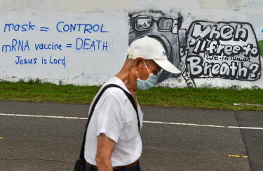 A man wearing a face masks walks past graffiti attacking face masks and vaccines, and proclaiming 'Jesus is Lord', sprayed on a wall along a bicycle path.