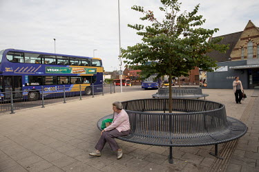 An elderly woman sits on a bench near the town bus station.
