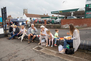Tourists sit on a wall near the sea front eating take away fish and chips.