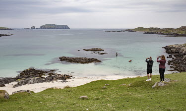 Tourists on the west coast of the Isle of Lewis taking photos as a kayak paddles towards the beach. The Isles of Harris and Lewis have much smaller numbers of visitors compared to more easily accessib...