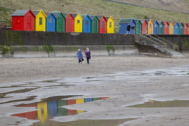 Two elderly ladies walk along the beach with colorful beach huts in the background.