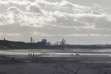 Three horseback riders and people walking on the beach at Saltburn by the Sea, Yorkshire, England with Redcar Steel works (now shut down and permanently decommissioned) in the background.