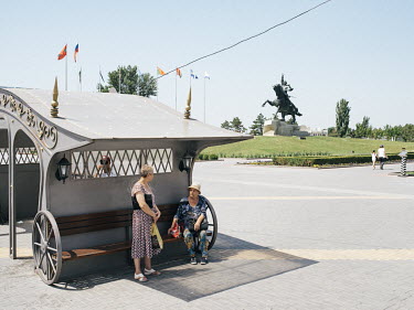 Two women wait at a public transport stop in the centre of Tiraspol. In the background, a statue of Alexander Suvorov, Russian count and general (1730 â�"1800).