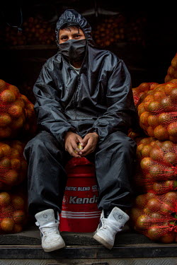 An orange seller wearing protective clothing and a face mask sits on an oil tin.