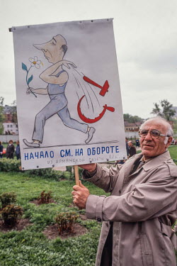 An anti Communist protester holds a hand made sign showing a man holding flowers and throwing away a hammer and sickle. The reverse shows an ape holding a hammer and sickle with the caption 'Totalitar...