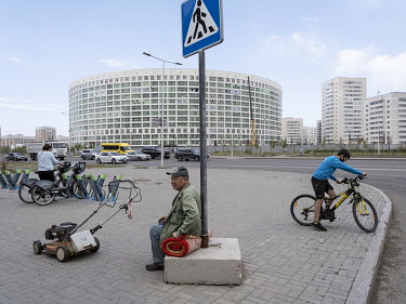 A man sits on a rug next to a lawnmower on a pavement in front of a circular apartment building in the eastern outskirts of the city.