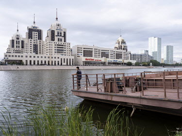 A man stands on an old boat on the Ishim river with the Radisson Hotel in the background.
