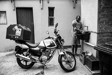 Paul, a food delivery rider, washes dishes outside his room next to his motorbike. He lives in a small room in a complex shared with several other drivers.