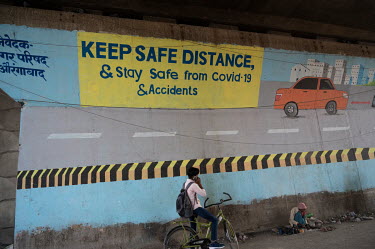 A boy on a bicycle speaks on a mobile phone whilst a cobbler wearing a pink hat repairs shoes beneath a mural about road safety and COVID-19.