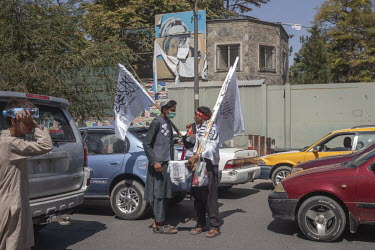 Two street vendors carrying Taliban flags chat amid traffic jams. In the background is a portrait of Commander Massoud and the French school, struck by a terrorist attack in 2014.