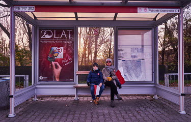 A woman and a boy holding Polish flags wait at a bus stop on National Independence Day.