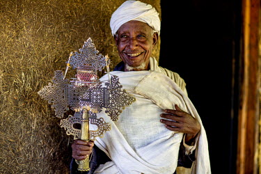 A smiling priest holds a large metal coptic cross at Debre Mariam church.