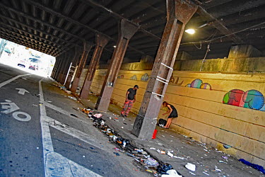 A couple stand beneath a bridge on a pavemnet littered with drug injecting paraphernalia in North Philadelphia, between Huntingdon and Allegheny on the elevated Market-Frankford subway. This is the ep...