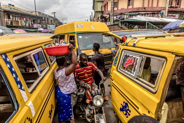 Pedestrians and motorcyclists navigating the narrow spaces between passenger vehicles at the Idumota Market on Lagos Island.
