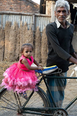 A grandfather pushes his granddaughter along on a bicycle. She is wearing a very bright pink dress.