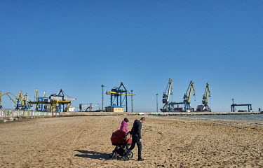 A couple pushing a baby pram (stroller) through the sand on a beach by the Azov Sea. In backgound are cranes at the Port of Mariupol.