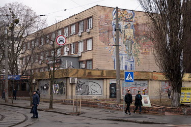Pedestrians walk along a street opposite a building decorated with a Soviet-era mosaic.