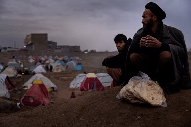 Two men squat on the ground above the tents in a makeshift camp where about one thousand people have moved, and are now living in subhuman conditions, from their villages and farms in the countryside...
