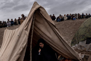 A woman sits in a makeshift tent while men from the camp are passing on their time on a small hillside.   About one thousand people have moved from their villages and farms in the countryside due to t...