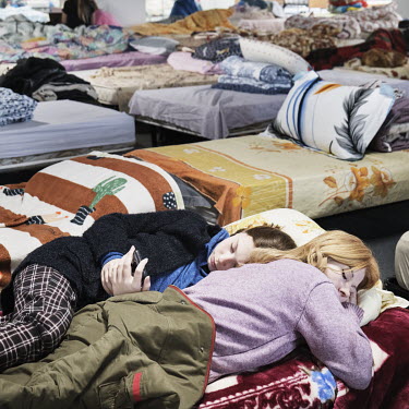 A girl lies next to her sleeping mother and looks at her phone in a temporary reception centre for Ukrainian refugees.