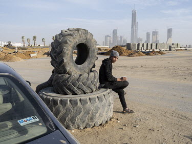 A worker sits on a stack of tyres and drinks tea in a roundabout in Egypt's New Administrative Capital. In the background are newly finished towers in the Central Business District.~Following persiste...