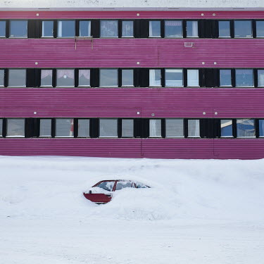 A car buried by a winter snow fall beside a pink building.