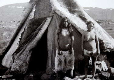 An old photograph of an Inuit couple standing outside a teepee.