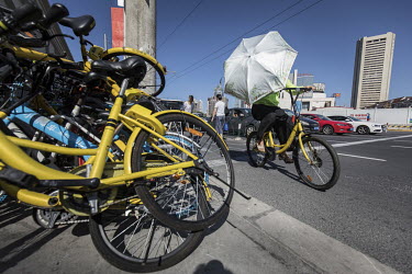 A cyclist, sheltering from the sun behind an umbrella, rides past a pile of shared bicycles from various providers. While cheap, convenient, and theoretically environmentally friendly, the craze and c...