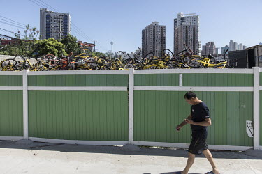 A pedestrian walks past a mountain of discarded shared bicycles piled up in a compound. While cheap, convenient, and theoretically environmentally friendly, the craze and commercialisation of shared b...