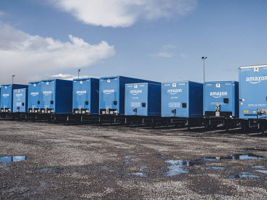 Amazon 'Prime' trailers parked on a gravel parking lot between the Rugeley 'BHX1' fulfilment centre and a construction supplies store.
