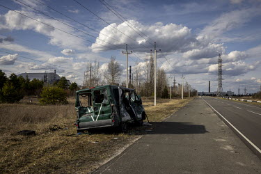 A destroyed vehicle, bearing the letter 'V', now synonymous with the Russian military, left on the side of the road leading to the Chernobyl power plant. In the background, is the New Safe Confinement...