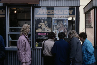 A group of women look at western movie and pip-up posters in a shop window display. They include a movie poster for Rambo III and a poster featuring 1980s glamour model and minor pop start Samantha Fo...
