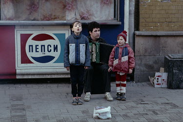 A father plays an accordian as his children sing for money while busking next to a Pepsi-Cola advertisement.
