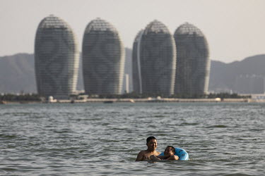 Two men swim in the sea while behind them can be seen high rise residential housing on Phoenix Island, an artificial archipelago developed by Sanya Phoenix Island Development Co.