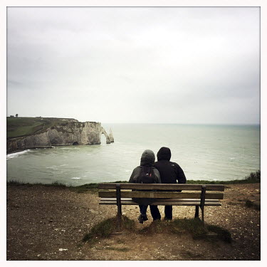 A couple sit on a bench looking towards the Etretat chalk cliffs, arches and stacks in the English Channel.