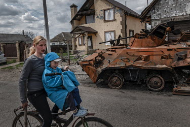 A woman and a child ride a bicycle past a destroyed tank.
