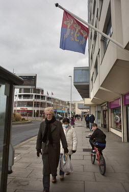 People walk past a building on the sea front flying a Hasting heraldic flag featuring the three lions/ships.