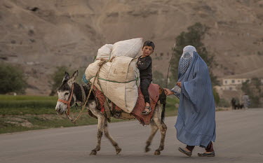A woman wearing a blue burqa walks with her child who rides on a donkey.