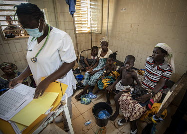 A member of the medical staff at the Kaya Regional Hospital reads the case notes for some children sitting with their guardians on a bed behind her. The hospital is operating at over 100% capacity, in...