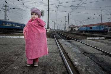 A child refugee, wrapped in a pink robe, stands beside the tracks at Lviv Central railway station.