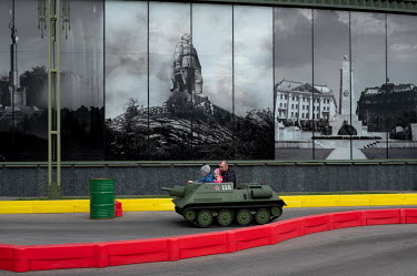 A father and child sit in a miniature tank in front of large photographs relating to the Soviet Union's military history in Patriot Park.