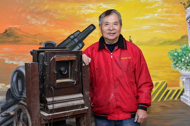 Retired photographer Mr. Chen Yi-pan stands beside his studio camera with his favourite hand-painted backdrop, in Tien Ao village. Chen used to make a good living taking portraits of the many ROC troo...