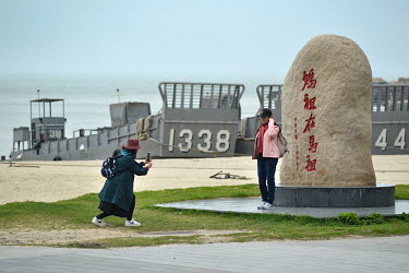 Domestic tourists from mainland Taiwan take photos at a stone monument with a backdrop of ROC Navy landing craft moored on the beach on Nangan Island. Nangan is the main island in the Matsu archipelag...