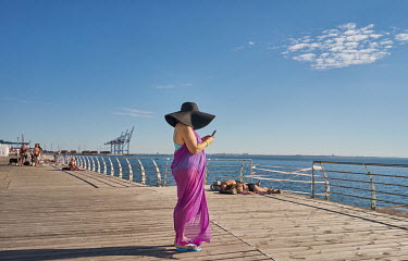 A woman wearing a large hat looking at smart phone on the Lanzheron Beach promenade where, despite Russia's war, people are sun bathing.