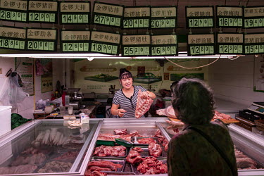 A woman sells pork at a wet market where various prices are displayed.