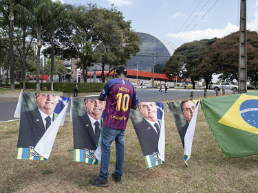 A street vendor, wearing a Messi football shirt, sells flags featuring president Jair Messias Bolsonaro. The building in the background is the Brasilia Shopping Mall.