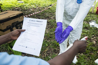 A scientist holds a list of instructions for donning and doffing ppe as they spray a colleague with disinfectant during an expedition, part of USAID Predict project, looking for new viruses in the wil...