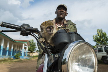 A farmer with a pet monkey riding on the mudguard of his motorcycle.
