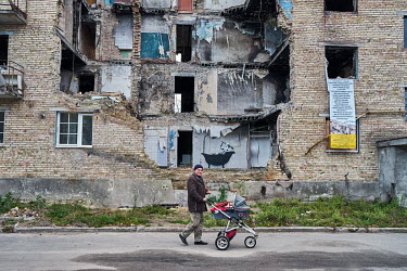 A man pushing a baby's pram (stroller) past a destroyed apartment block as life slowly returns to the village of Horenka. A mural of a man in a bath has been painted on the wall of an apartment expose...