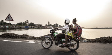 A motorcyclist carrying a woman holding a toddler as they ride along a coastal road.As motorcycle ownership in sub-Saharan Africa has grown from less than five million in 2010 to an estimated 27 milli...